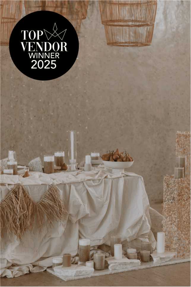 Whispering Thorns Top Vendor Winner 2025 – Award Winning Table Decor A styled Whispering Thorns all-inclusive wedding sweetheart table featuring draped natural linen, abundant white pillar candles, and a minimalist bowl of pears, overlaid with the 'Top Vendor Winner 2025' logo against a raw concrete wall and woven basket lighting.