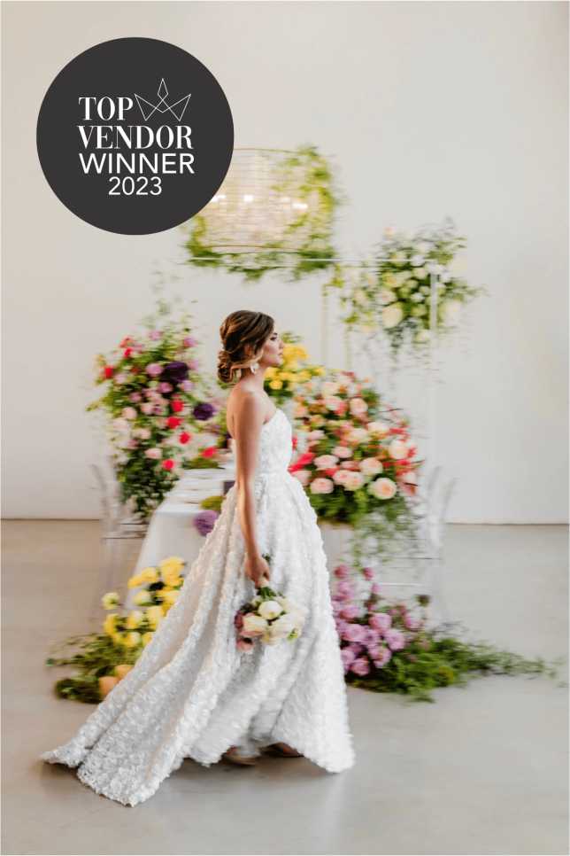 Whispering Thorns Top Vendor Winner 2025 – Award Winning Table Decor A styled Whispering Thorns all-inclusive wedding sweetheart table featuring draped natural linen, abundant white pillar candles, and a minimalist bowl of pears, overlaid with the 'Top Vendor Winner 2025' logo against a raw concrete wall and woven basket lighting. Premier glass wedding venue