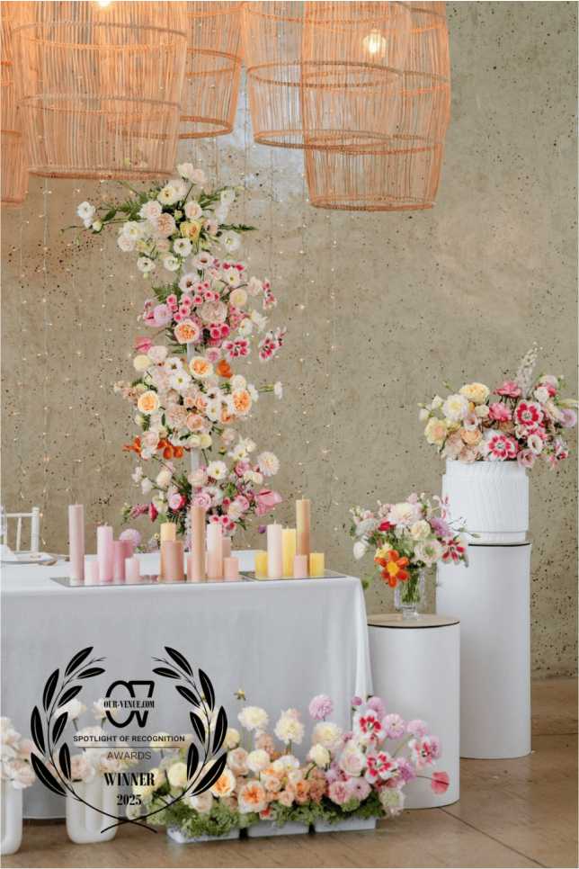 whispering-thorns-all-inclusive-wedding-decor-2025-winner A styled wedding reception setup at Whispering Thorns glass venue featuring abundant pastel floral arrangements, a column of pastel taper candles, modern white cylindrical plinths, and unique hanging wicker basket pendant lights. The Our-Venue.com 2025 winner award is overlaid against a raw concrete wall and fairy light backdrop.